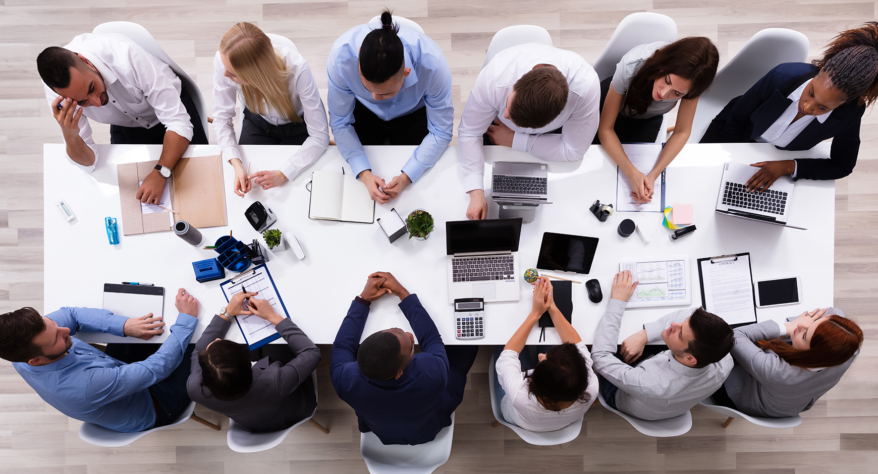 Overhead shot of 12 people working at a table filled with various work-related peripheral material: notebooks, clipboards, laptops, calculators, post it notes...