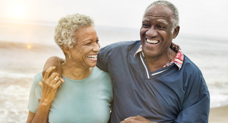 Smiling elderly black couple on a beach. The man's arm is around the woman's shoulder.